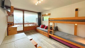 A room with bunk beds, a desk, a television, and a large window offering a view of buildings and mountains. - Economical studio for 4 with south-facing balcony in Les Menuires, pets allowed (Les Menuires)
