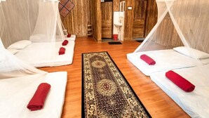 Una habitación con cuatro camas, cada una con mosquitero y una toalla enrollada. El suelo es de madera y hay una alfombra estampada.