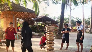 Un grupo de personas jugando Jenga en un espacio al aire libre, con una estructura de techo de paja y palmeras al fondo.
