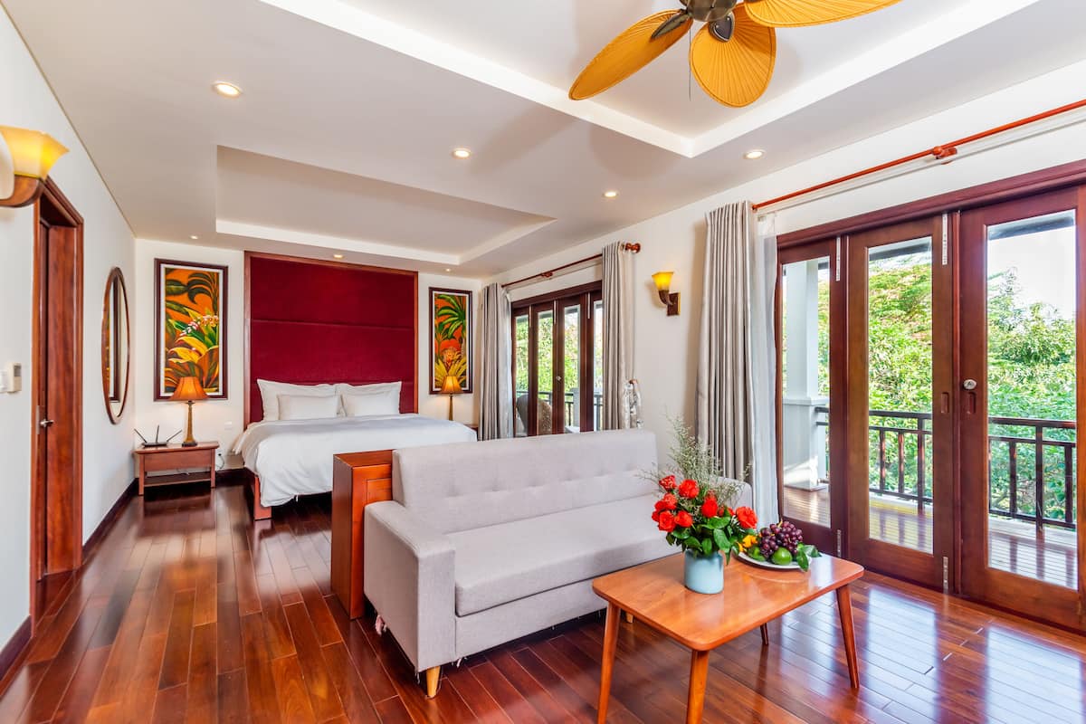 Habitación de hotel con cama, sofá, mesa de madera con un florero y balcón con vistas a la vegetación.