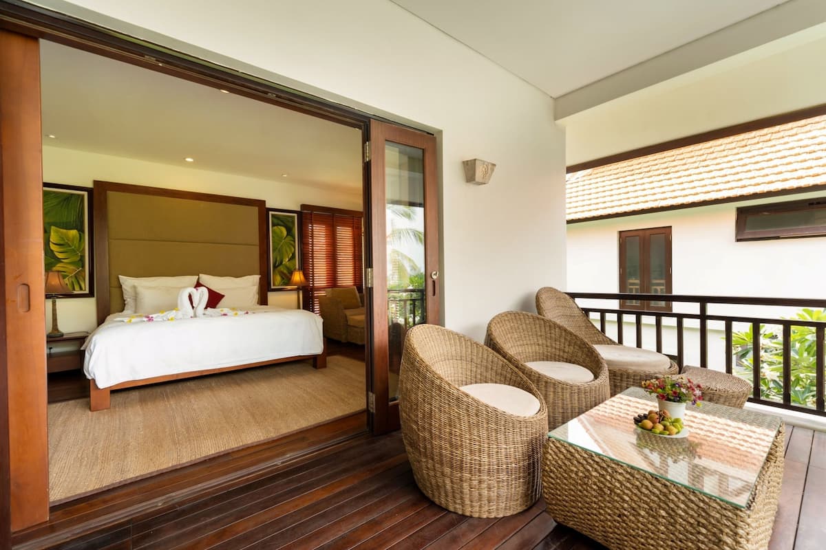Una habitación de hotel con una cama, un balcón con mobiliario de mimbre y vistas al verdor.