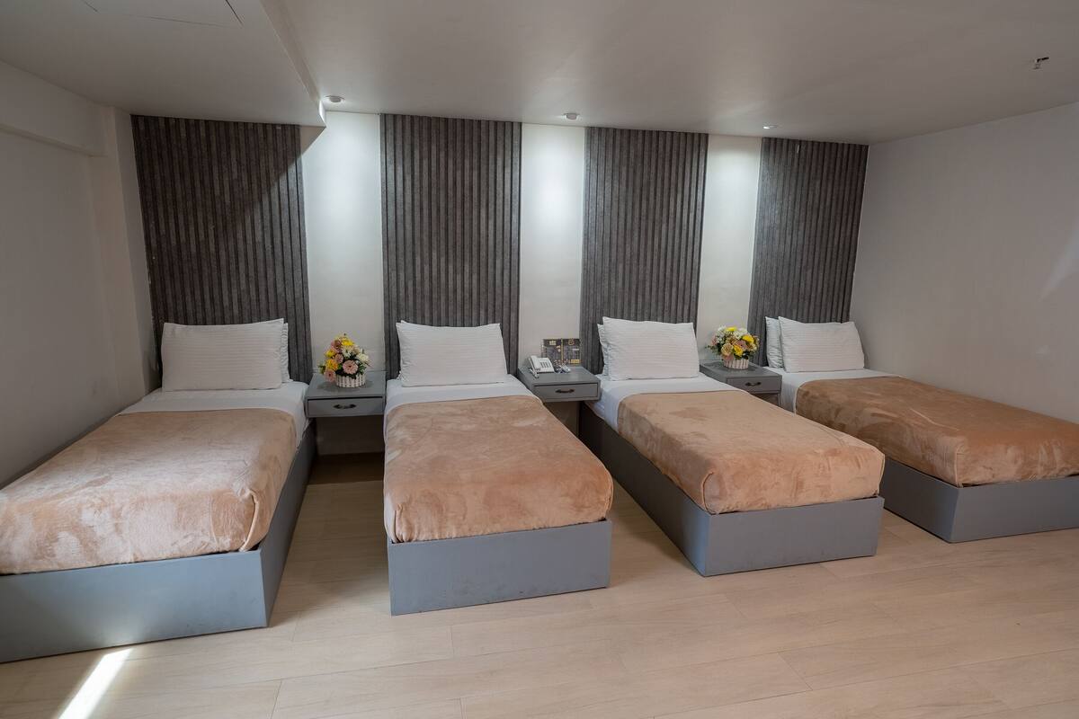 A hotel room with three single beds, each with a grey frame and a brown blanket. The room has a modern design with grey curtains and a wooden panel wall.