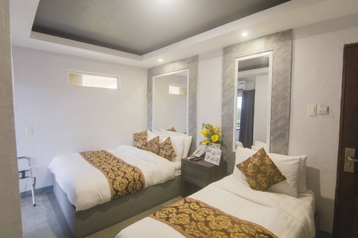 A hotel room with two beds, a small window, a mirror, a nightstand with a phone and a magazine, and a wheelchair near the window.