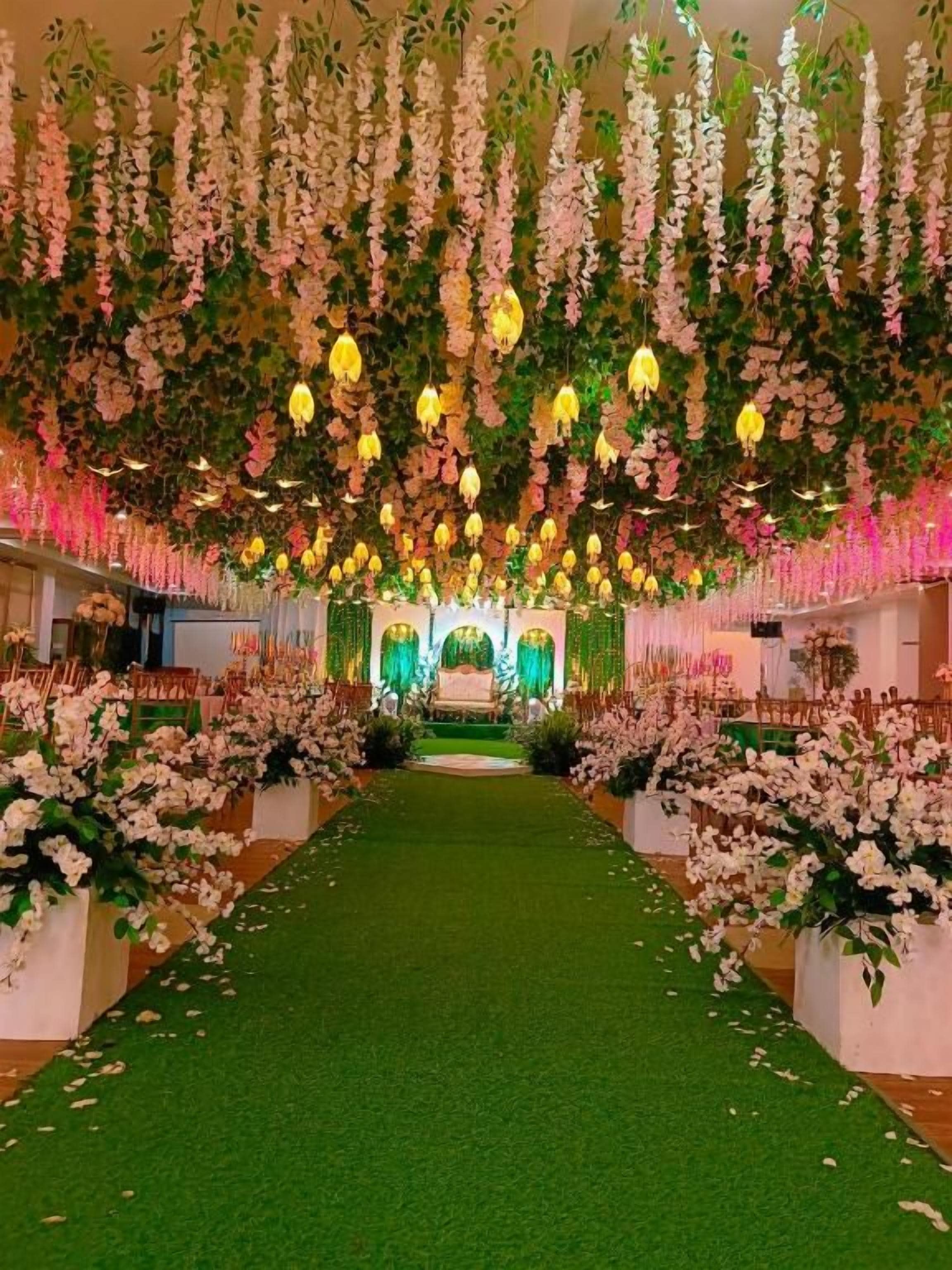 A venue with a central aisle leading to a stage, surrounded by floral arrangements and greenery.