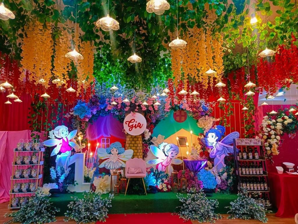 A colorful stage with fairy-themed decorations, including a sign that reads 'Gia,' and a backdrop with hanging plants and lights.