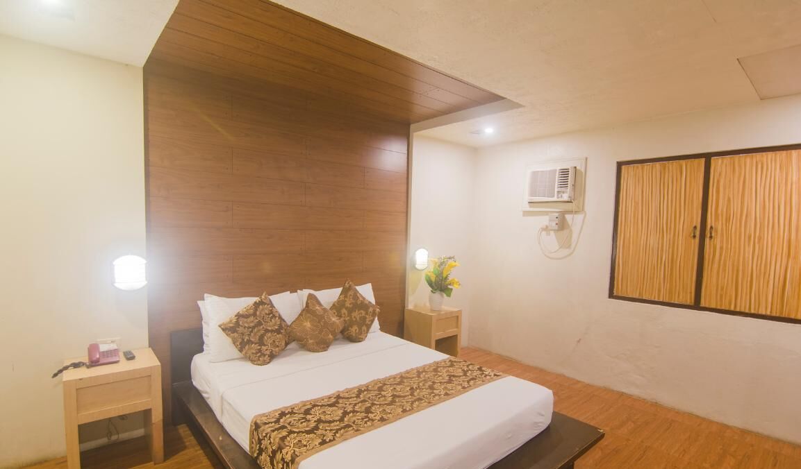 Habitación de hotel con cabecera de madera, cama con ropa blanca y almohadas estampadas, mesita de noche con lámpara y armario de dos puertas.