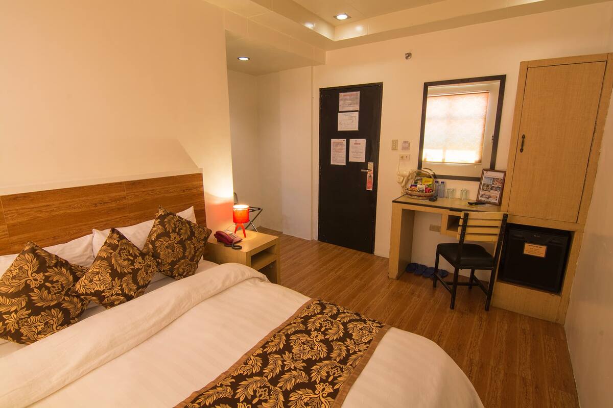 A hotel room with a bed, bedside table with a lamp, a desk with a chair, and a wardrobe.