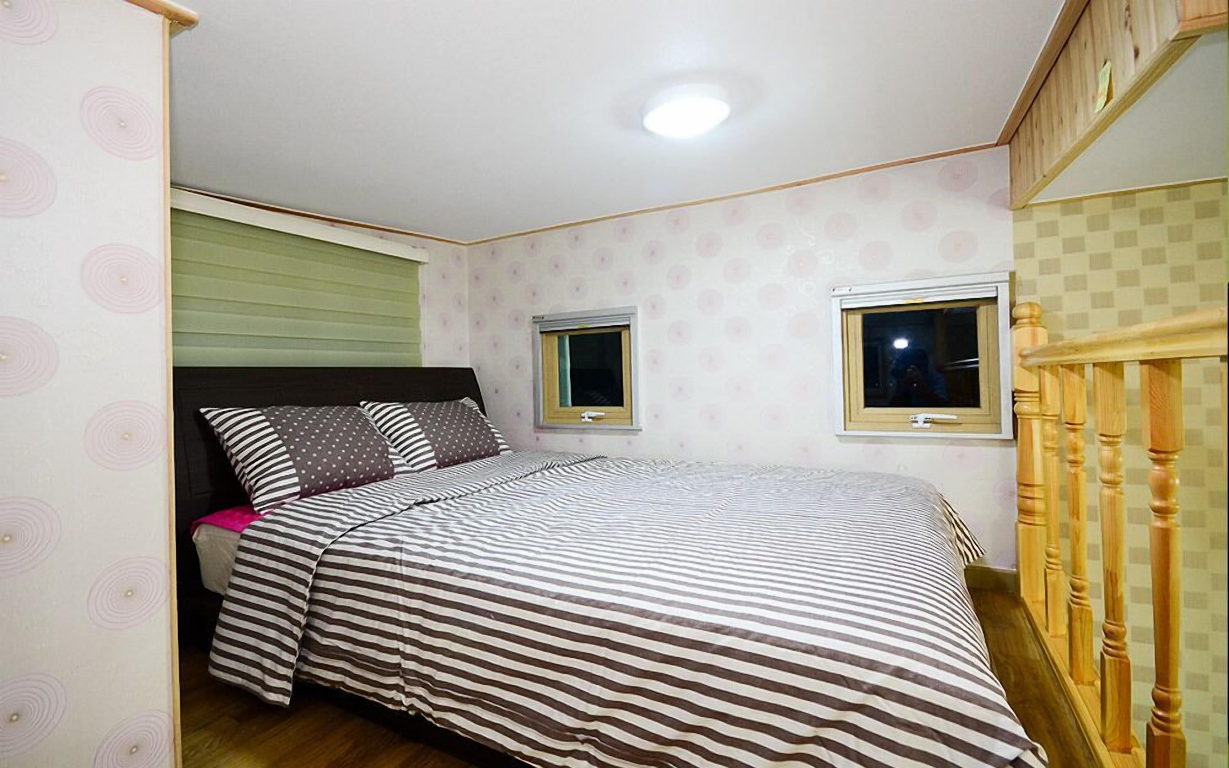 Quarto com colcha listrada, duas janelas, cabeceira de madeira e uma luminária no teto.