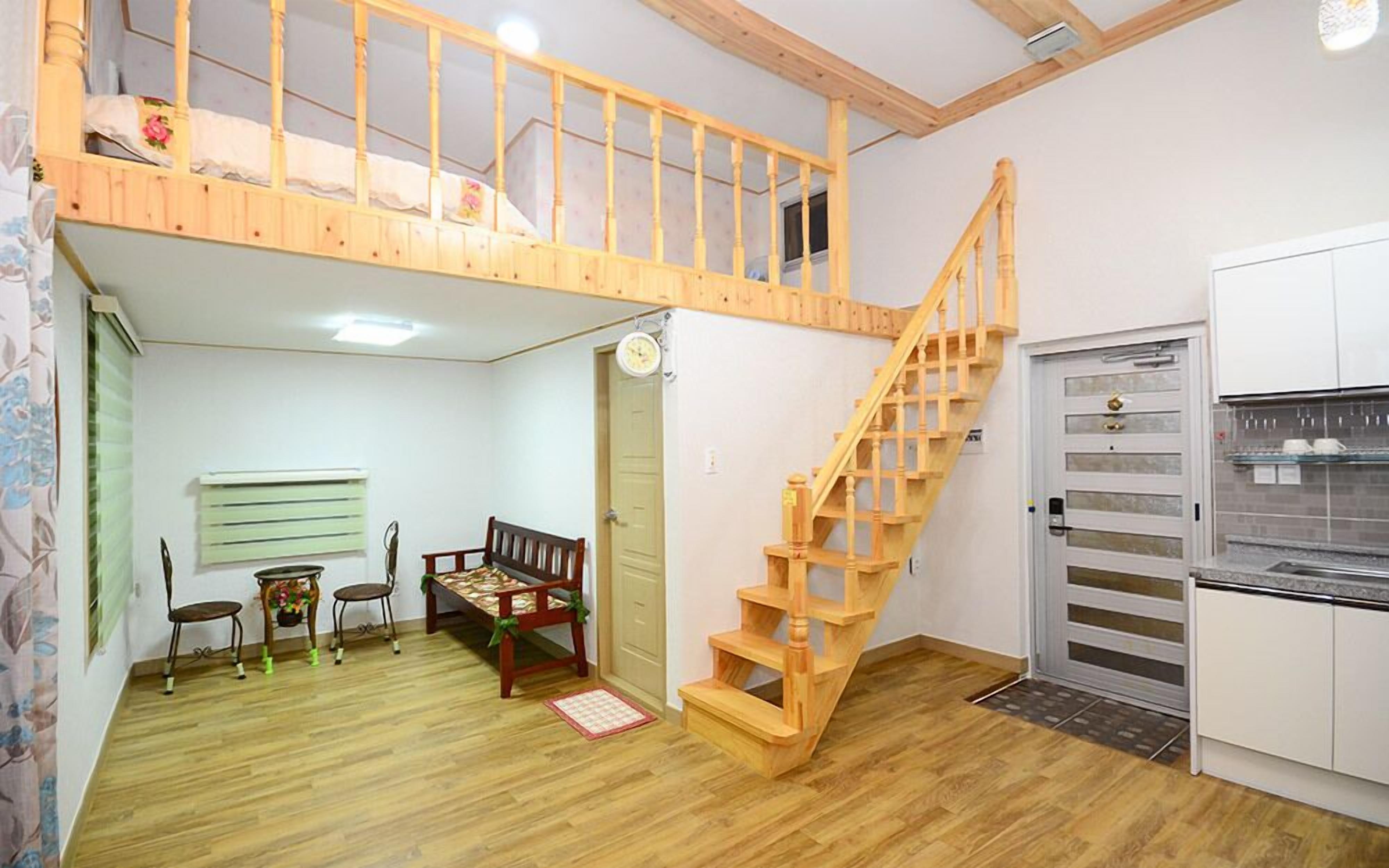 A room with a loft bed, a wooden staircase, a dining area with a table and chairs, and a kitchen with a refrigerator.