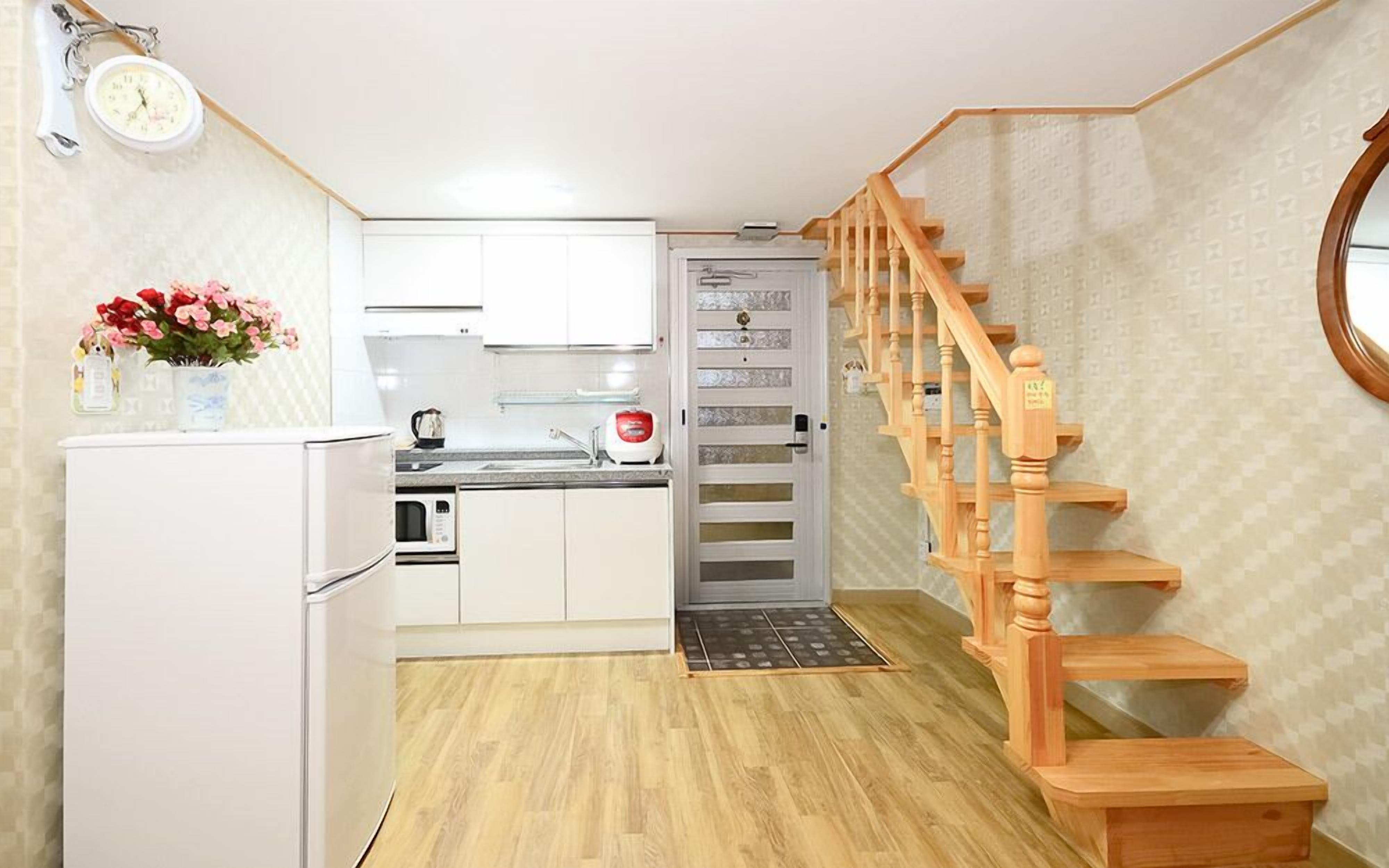 Een compacte keuken met witte kastjes, een magnetron en een koffiezetapparaat. Een houten trap met een leuning leidt naar een deur.