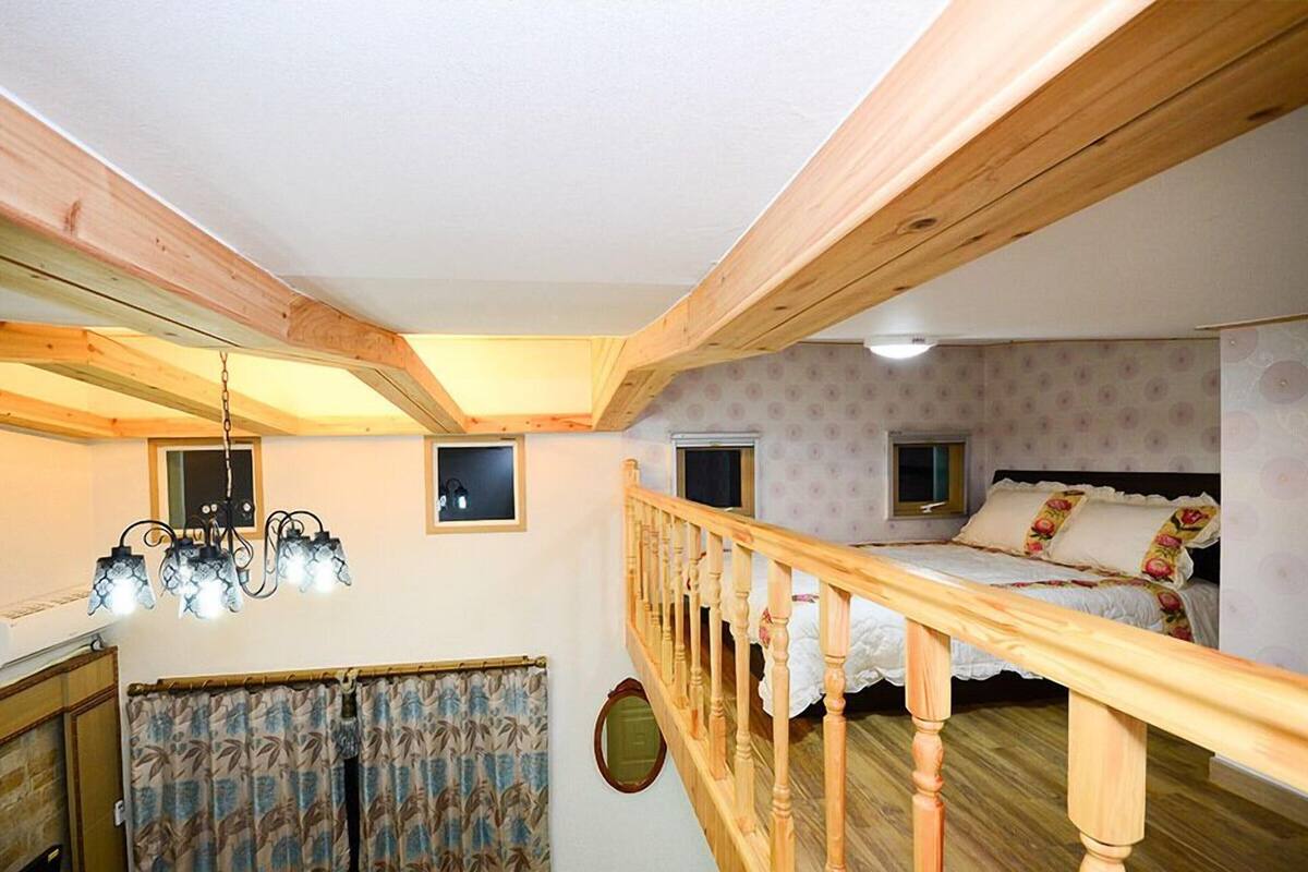 A bedroom with a wooden ceiling, a bed with floral bedding, a window, and a staircase with a wooden railing.