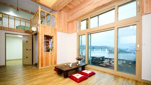 A spacious room with wooden floors, a wooden staircase, a dining area with a table and benches, and large sliding glass doors offering a view of the outdoors. - Sun and Moon Pension (Yeosu)