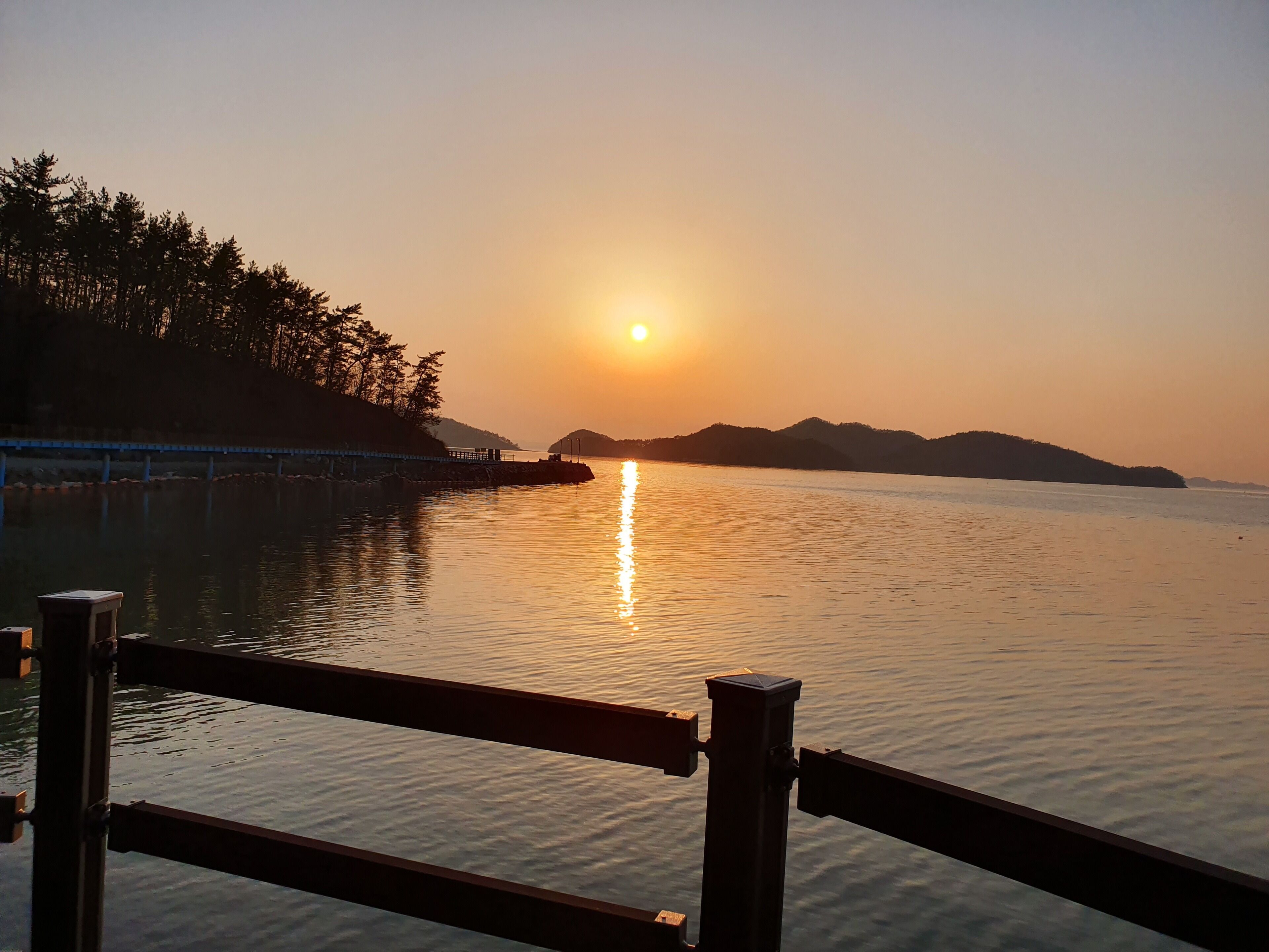 A sunset over a calm body of water with a pier and trees in the background.