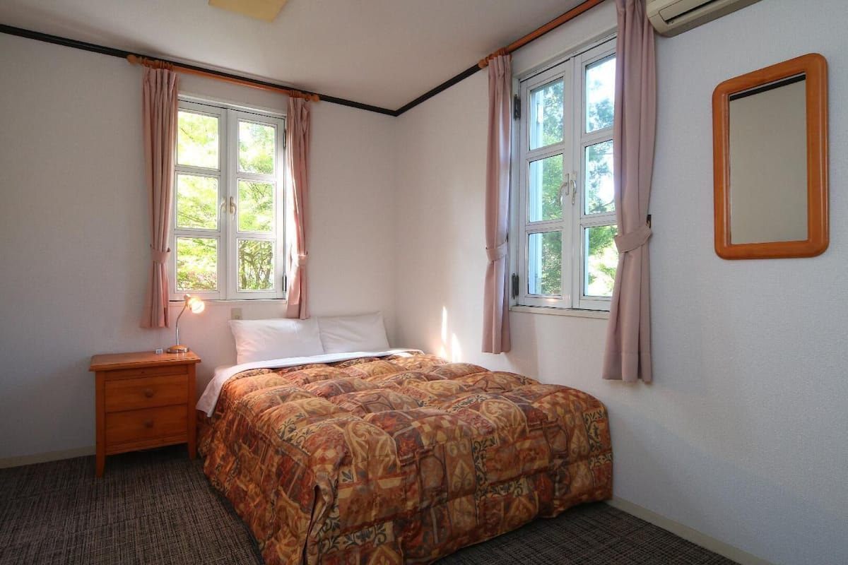 A bedroom with a bed, a nightstand, a mirror, and two windows with curtains.