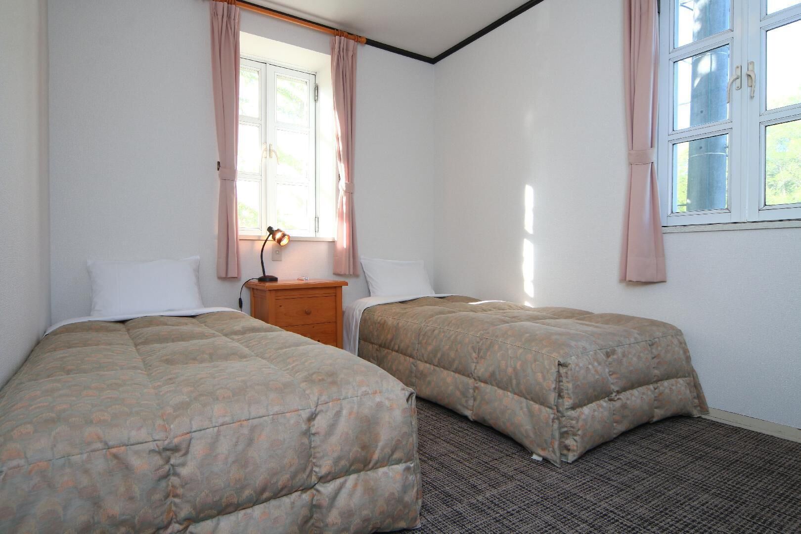 A room with two beds, a nightstand, a lamp, and a window with curtains.