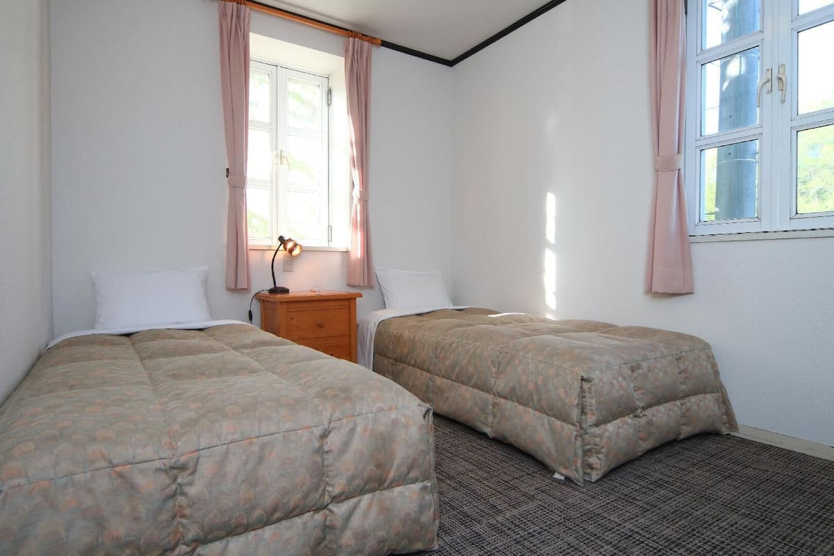 A room with two beds, a nightstand, a lamp, and a window with curtains.