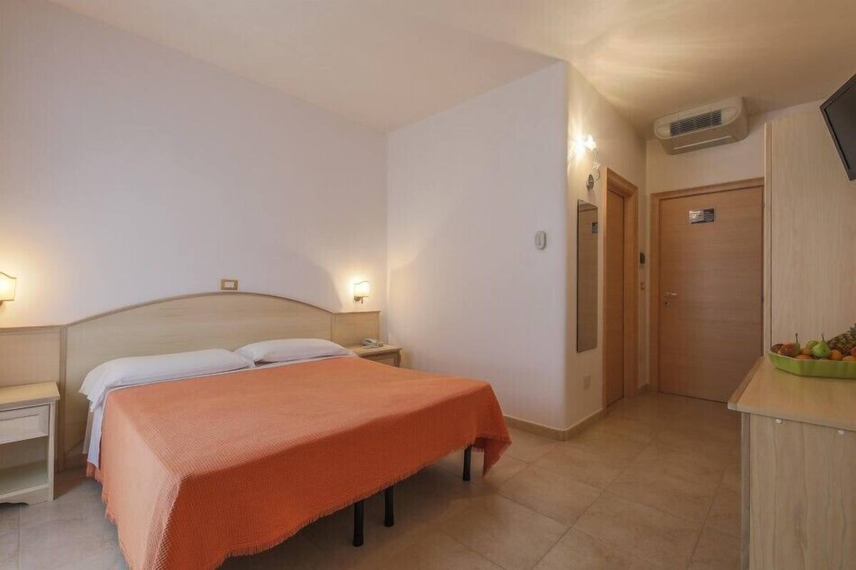 A hotel room with a bed, bedside tables, a nightstand, an air conditioning unit, and a small kitchenette with a fruit bowl.