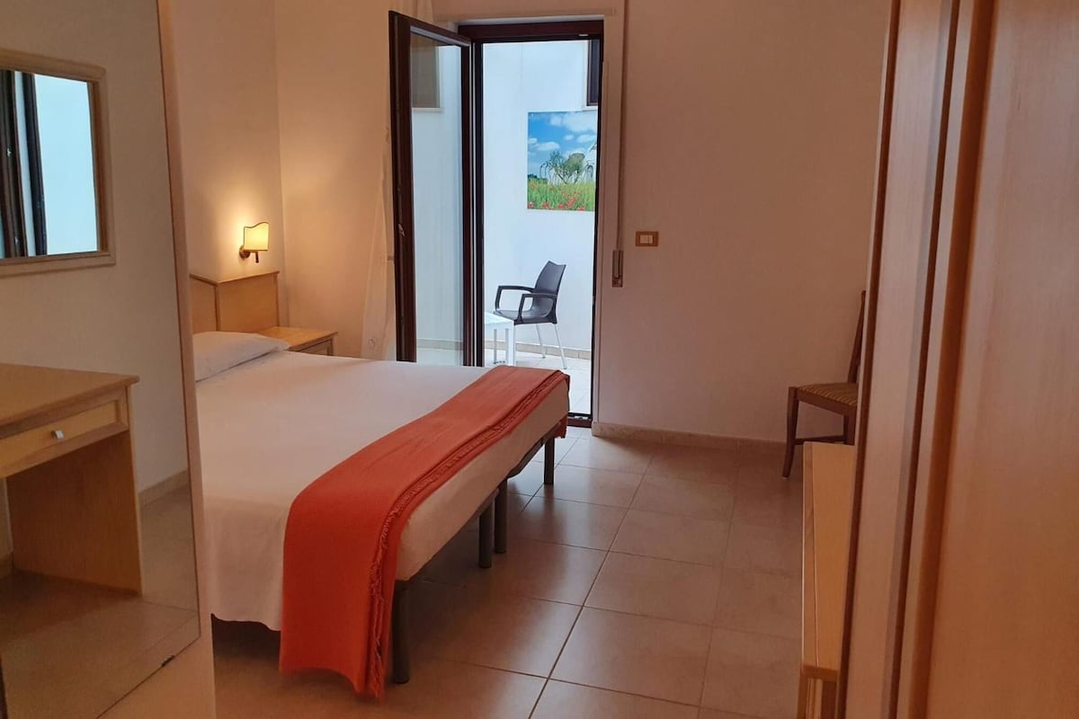 A hotel room with a bed, a desk, a chair, and a view of the outdoors through a glass door.