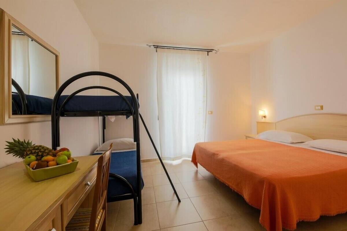 A room with bunk beds, a desk with a fruit bowl, a chair, and a bed with an orange bedspread.