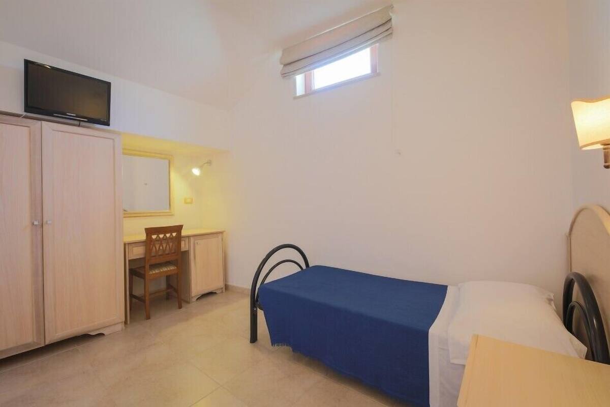 A small, well-lit room with a single bed, a desk, a chair, a television, and a window.