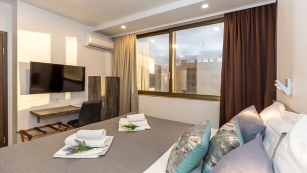 A modern hotel room with a large bed, a flat-screen TV, a desk, and a large window with curtains. - Apinelo Tower Rooms (Split)