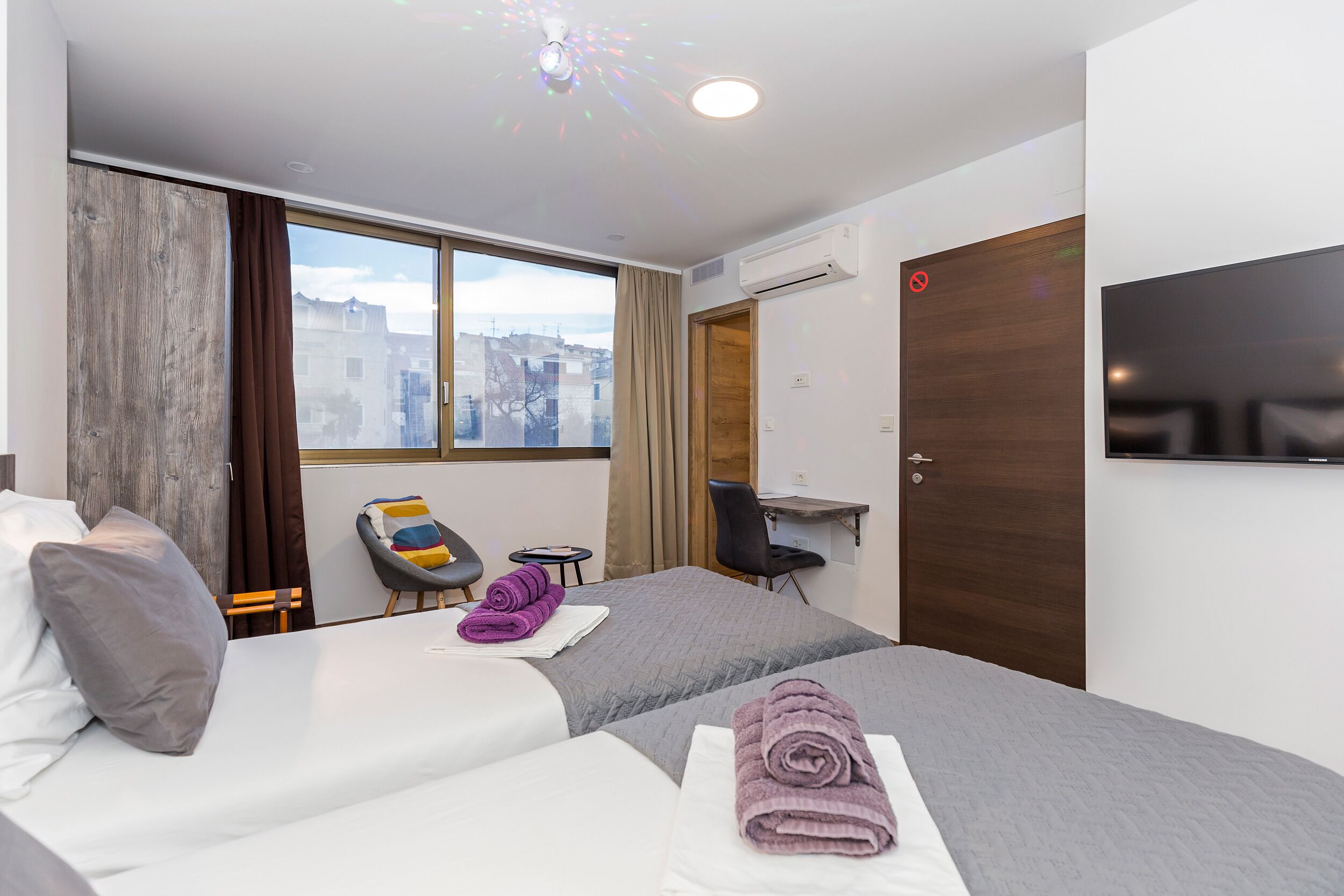 A hotel room with two beds, a desk, a chair, and a large window offering a city view.