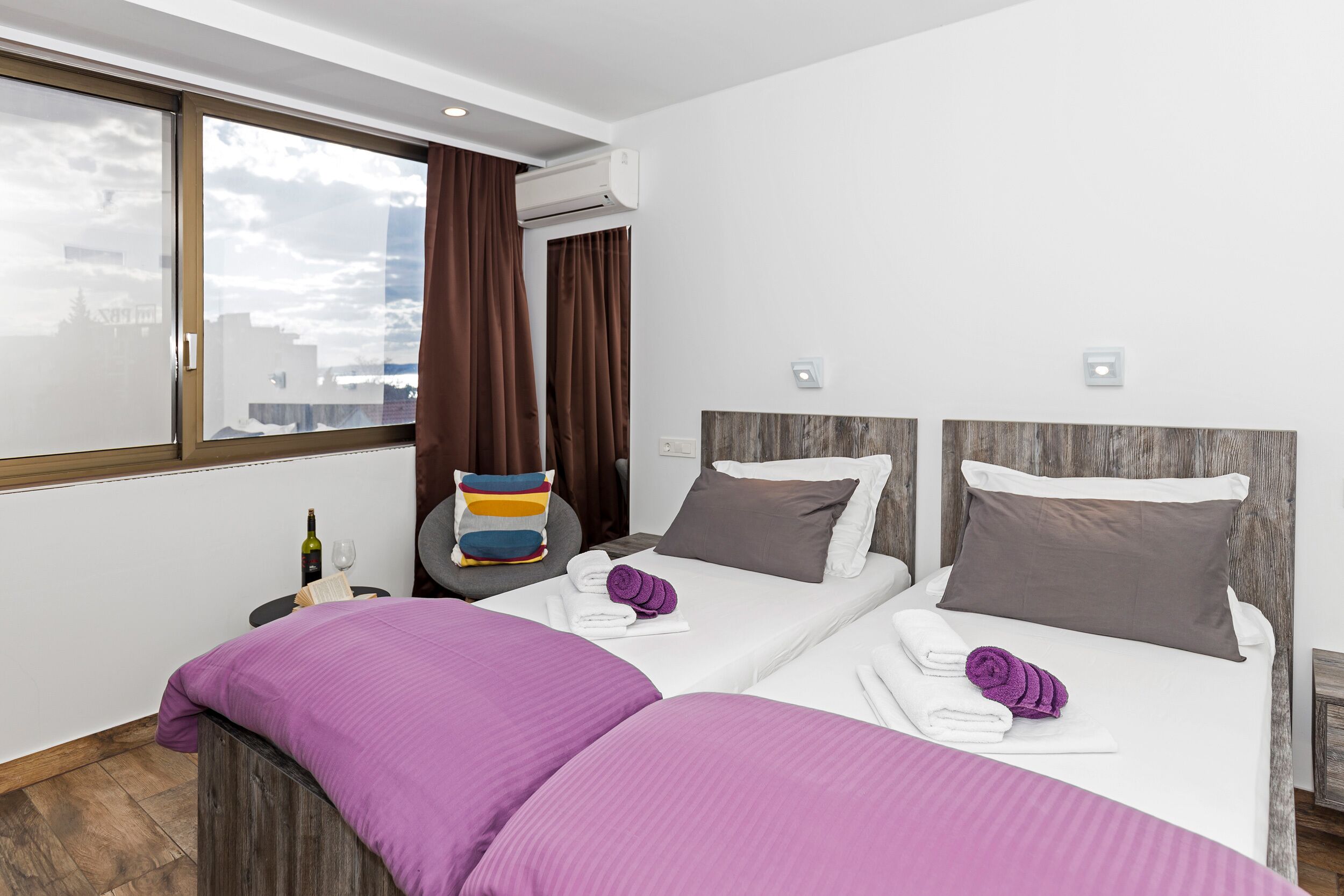 A hotel room with two beds, a window with curtains, an air conditioning unit, and a small table with a bottle and glass.