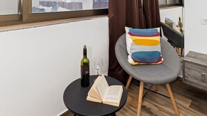 A modern living room with a large window, a cozy seating area, a small table with a book and a bottle, and a grey sofa with colorful cushions. - Apinelo Tower Rooms (Split)