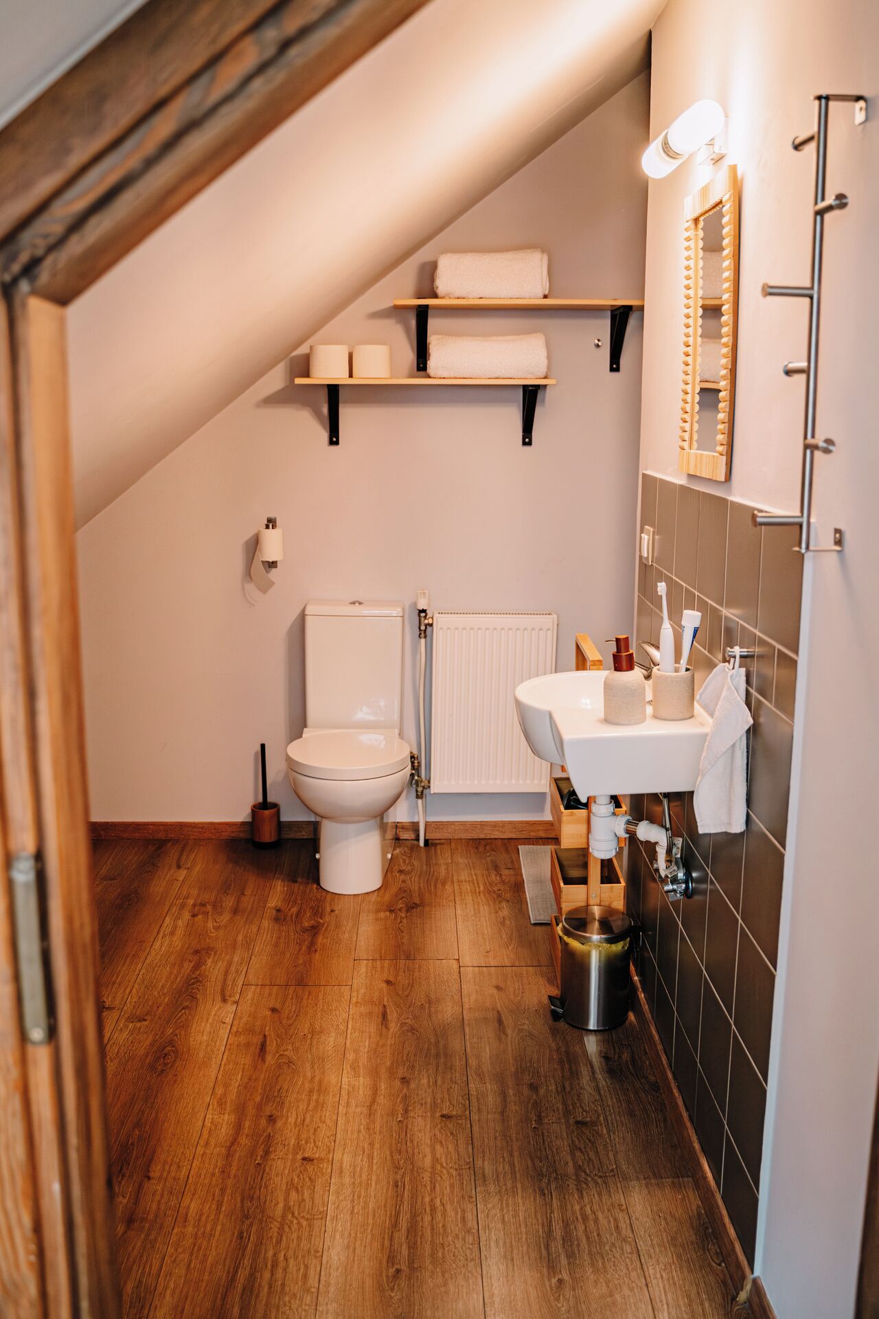 A small, cozy bathroom with a wooden floor, a white toilet, a sink with a mirror, and shelves above the toilet holding towels.