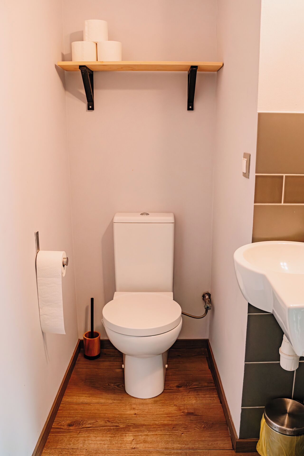 Ein kleines, kompaktes Badezimmer mit Toilette, einem Holzregal darüber, einer Toilettenpapierrolle und einem Metall-Abfalleimer.