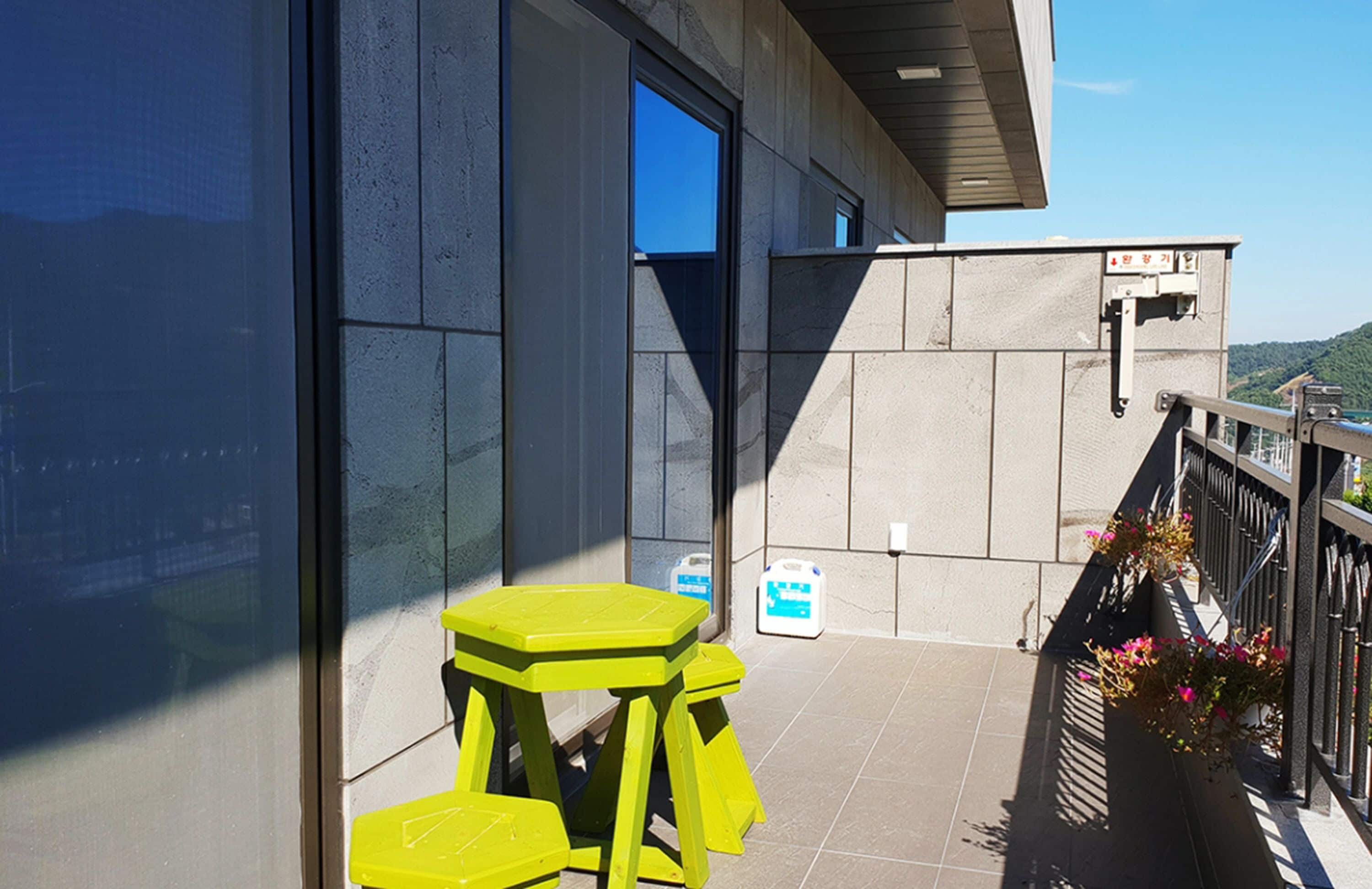 A balcony with a yellow table and chairs, a window, and a view of a building and hills.