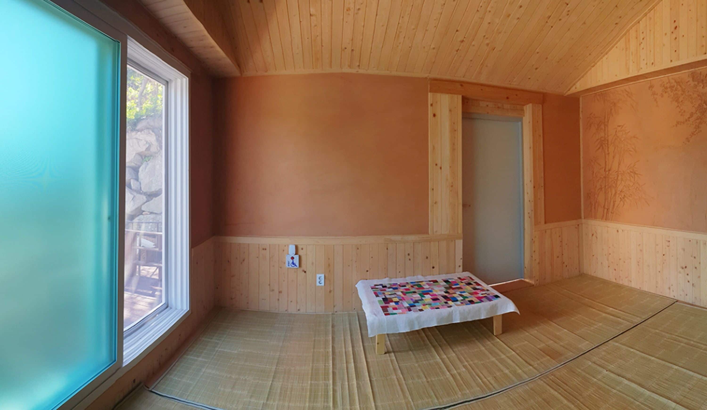 Una habitación con paredes y suelo de madera, una ventana, una puerta y un colcha en el suelo.