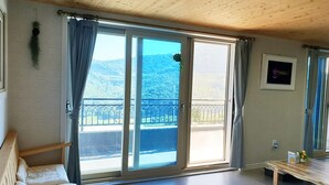 Ein Zimmer mit Holzboden, einer Glastür zum Balkon, einem kleinen Holztisch und einem Bett mit weißer Matratze.