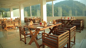 A spacious dining area with wooden tables and chairs, large windows offering a view of a forested mountain. - Dream Coconut Villa Resort (Devikolam)