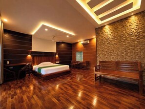 A hotel room with a wooden floor, a bed with a green bedspread, a desk, a chair, a bench, and a textured wall. - Dream Coconut Villa Resort (Devikolam)