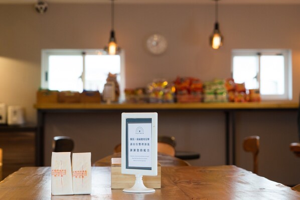 A wooden table with a menu stand displaying text, two white paper bags, and a kitchen area with hanging lights and food items in the background. - V Hotel (Taipei)