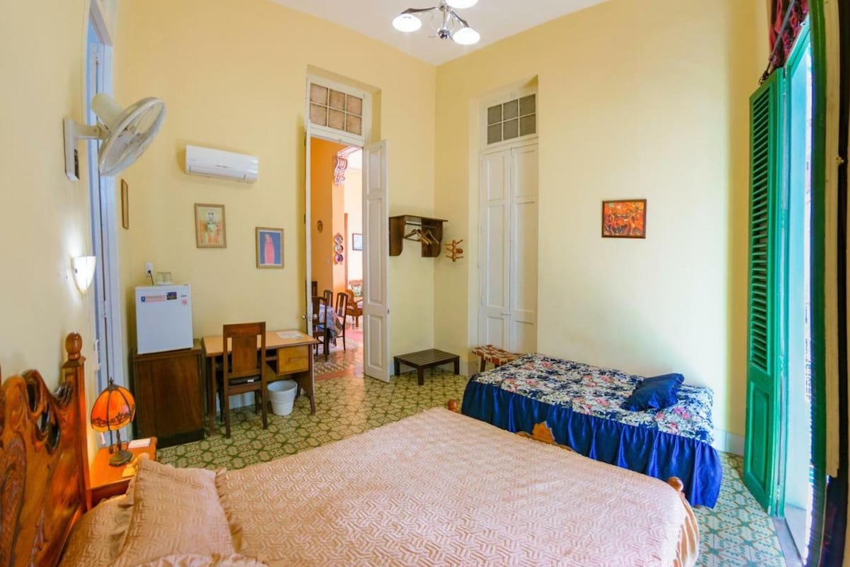 A bedroom with a bed, a desk, a chair, a fan, and a window with green shutters.