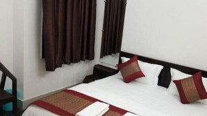 Une chambre d’hôtel avec un lit, deux oreillers, une couvre-lit rouge et marron, une couverture blanche, une table de chevet en bois, un miroir et des rideaux marron.