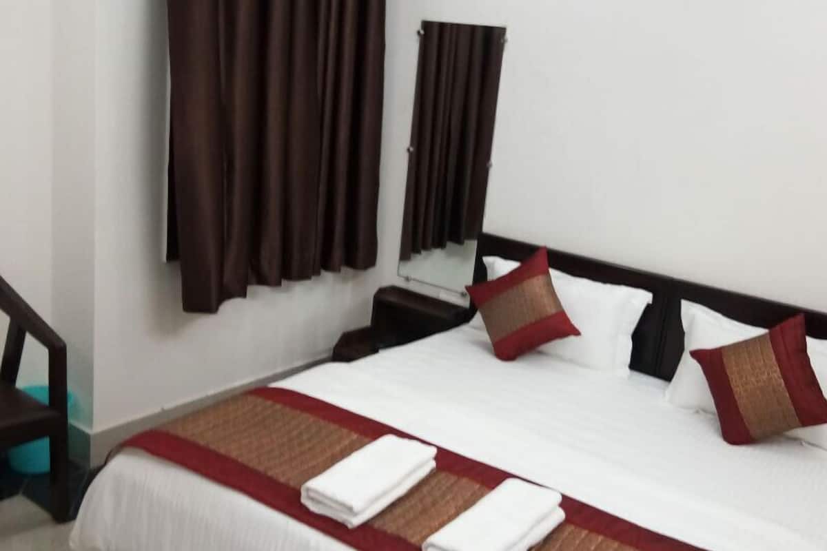 Une chambre d’hôtel avec un lit, deux oreillers, une couvre-lit rouge et marron, une couverture blanche, une table de chevet en bois, un miroir et des rideaux marron.