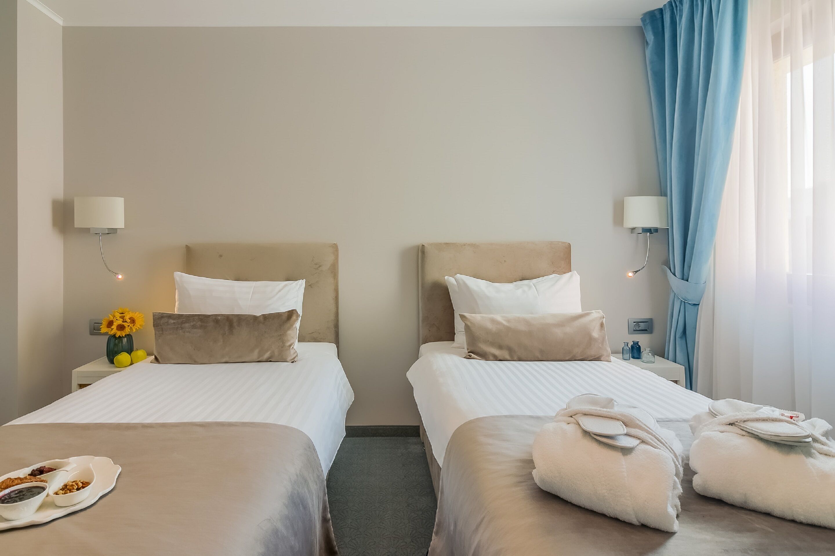 Habitación de hotel con dos camas, una mesita de noche con lámpara, un jarrón con flores y vistas al exterior a través de una ventana con cortinas.
