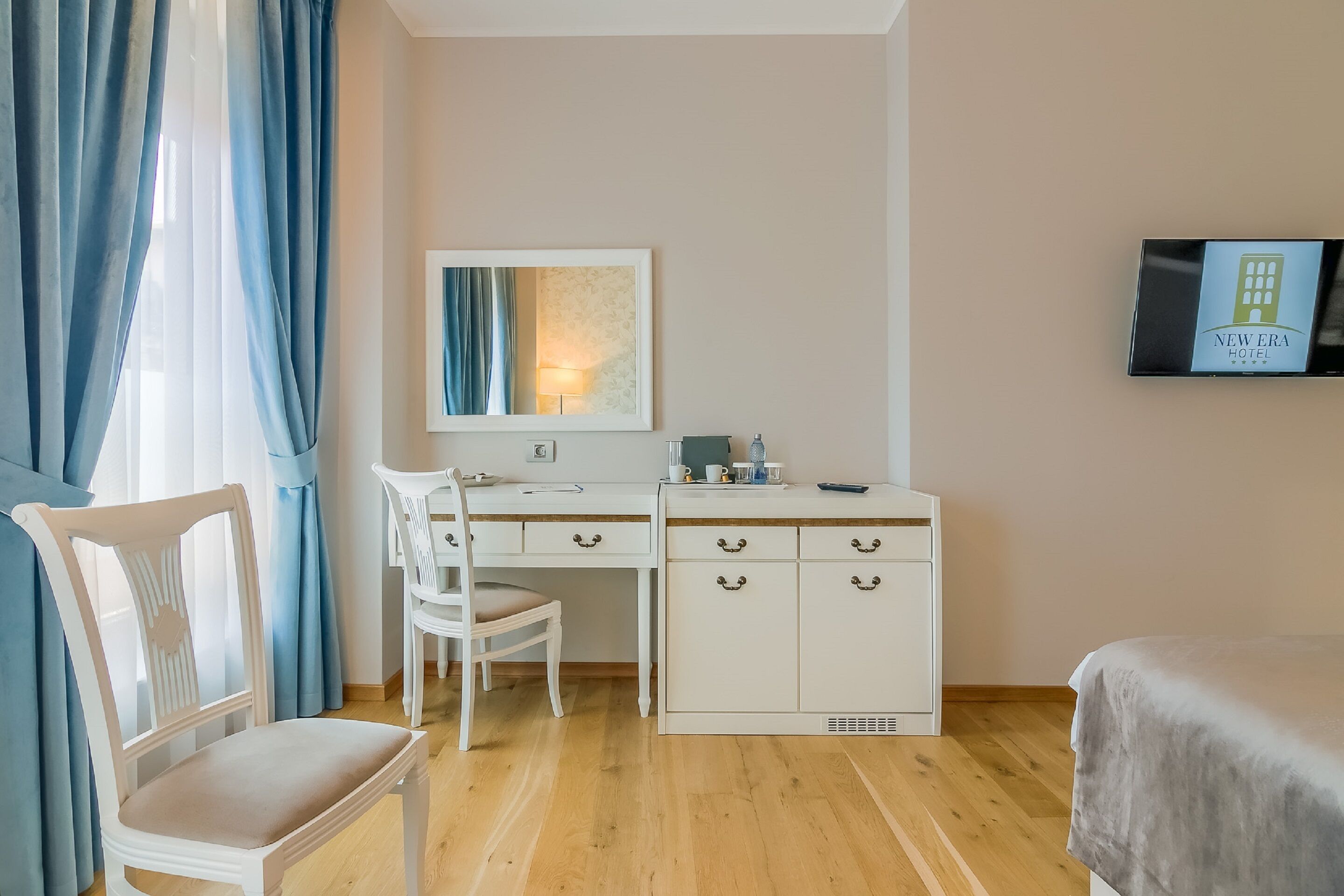 Quarto de hotel com piso de madeira, uma escrivaninha com cadeira, uma cama, televisão e um quadro na parede.