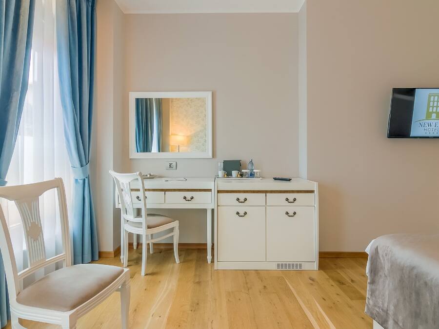 Quarto de hotel com piso de madeira, uma escrivaninha com cadeira, uma cama, televisão e um quadro na parede.