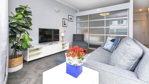 Ein modernes Wohnzimmer mit einem grauen Sofa, einem weißen Couchtisch, einem Flachbildfernseher, einer großen Kühlpflanze und einer Glastür zum Balkon.