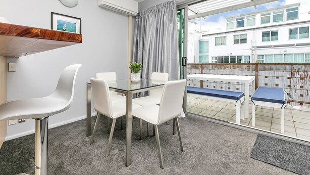 Ein modernes Zimmer mit einem Glastisch und weißen Stühlen, einer Topfpflanze und Blick auf ein Gebäude durch ein großes Fenster.