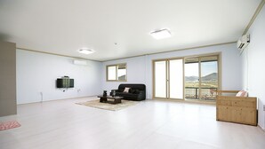 A spacious living room with a black sofa, a coffee table, and a TV mounted on the wall. There is a large sliding glass door leading to a balcony with a view of the outdoors. - Golden Field Pension (Yeosu)