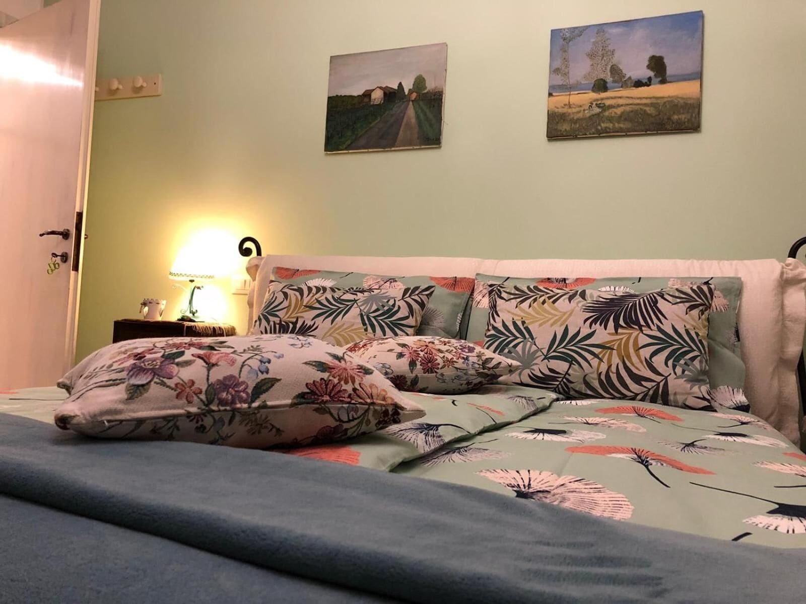 A bedroom with a bed, floral-patterned bedding, a bedside lamp, and two landscape paintings on the wall.