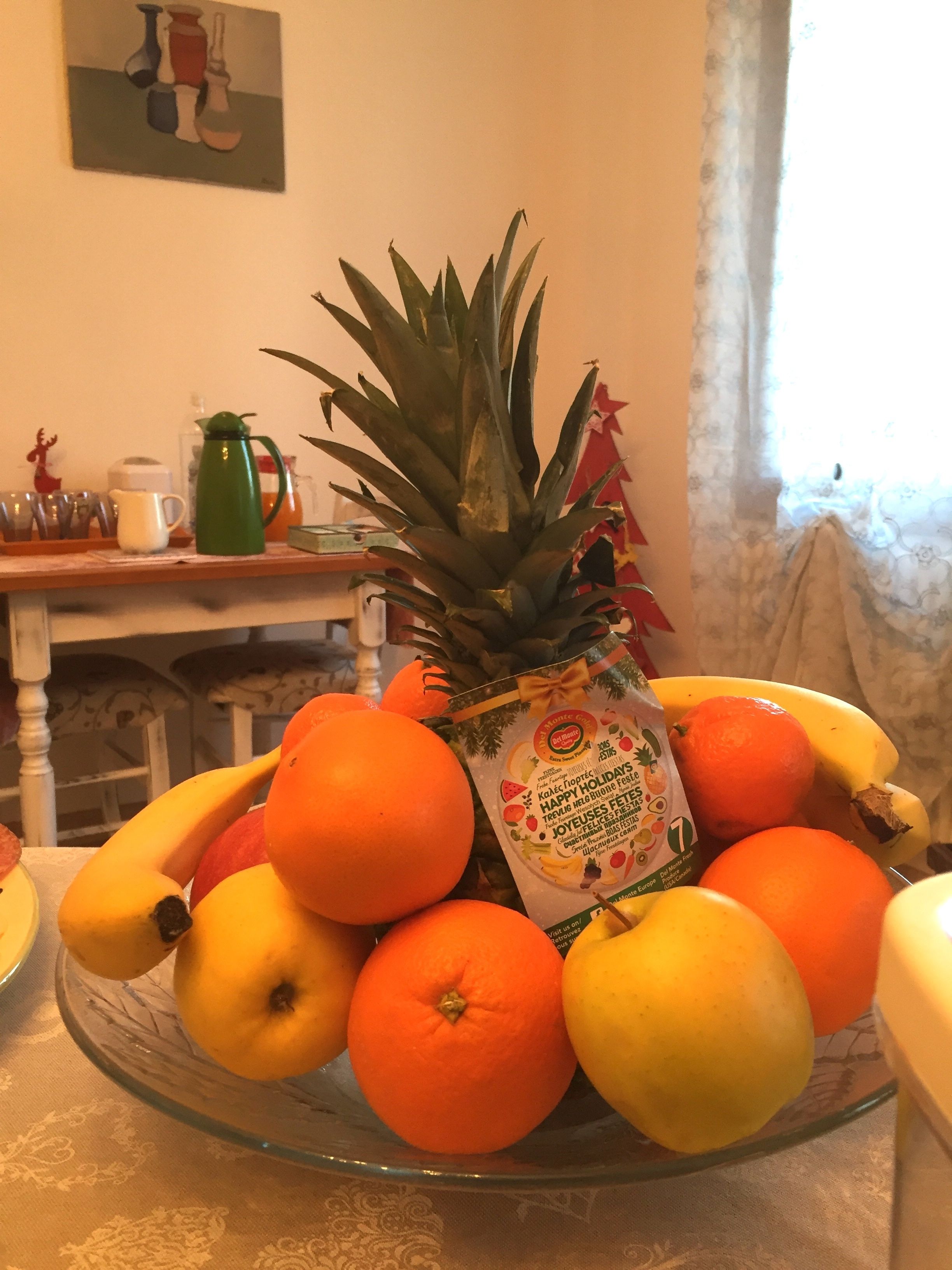 A glass bowl filled with fruits including bananas, oranges, and a pineapple, placed on a table with a patterned tablecloth.