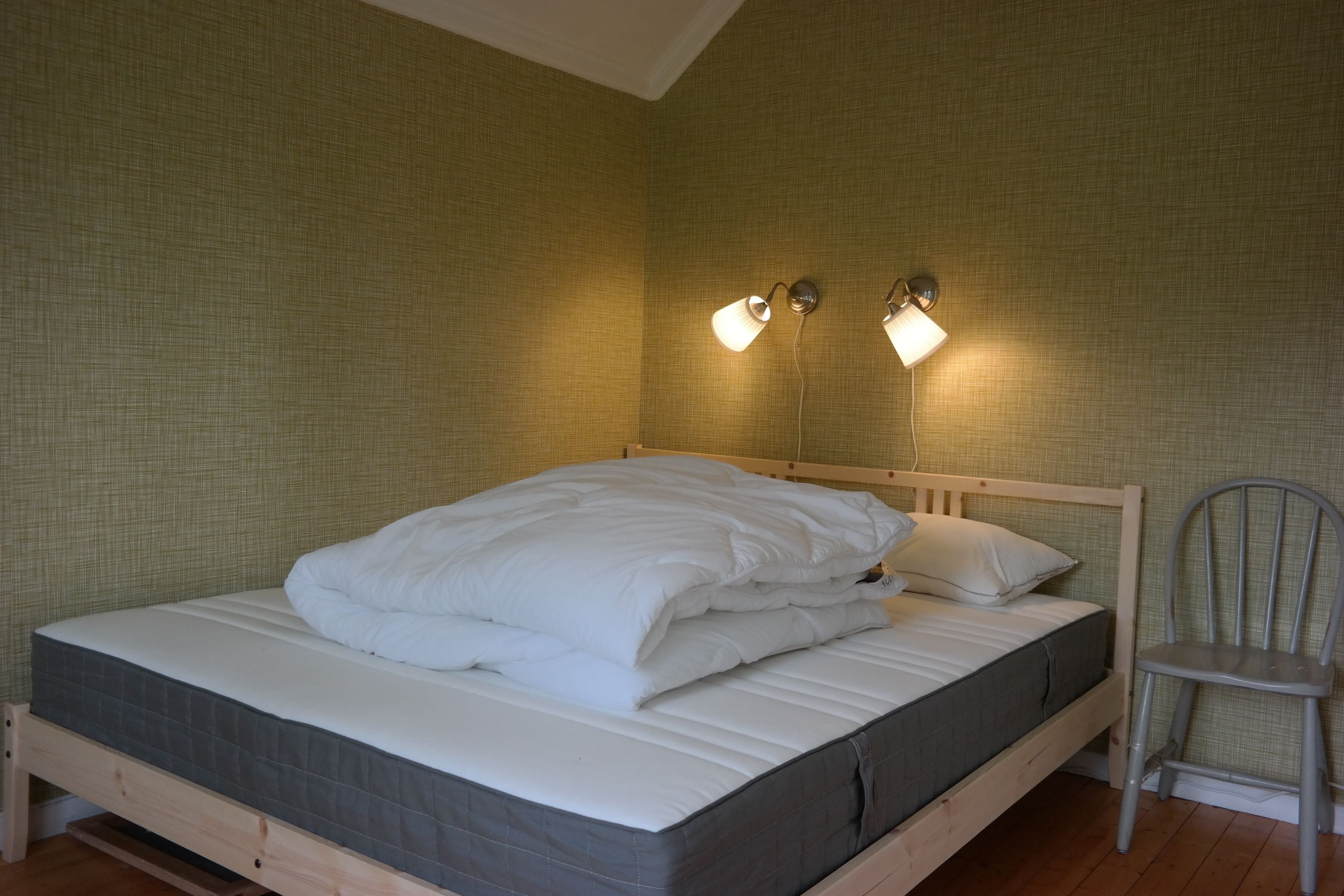 Una cama bien hecha con colchón gris y ropa de cama blanca, cabecera de madera y dos lámparas fijadas a una pared con textura.