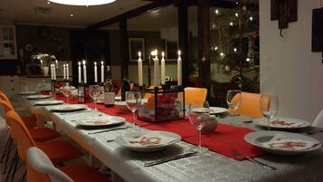 Een eetgedeelte met een lange tafel gedekt voor een maaltijd, kaarsen en een kerstboom op de achtergrond.