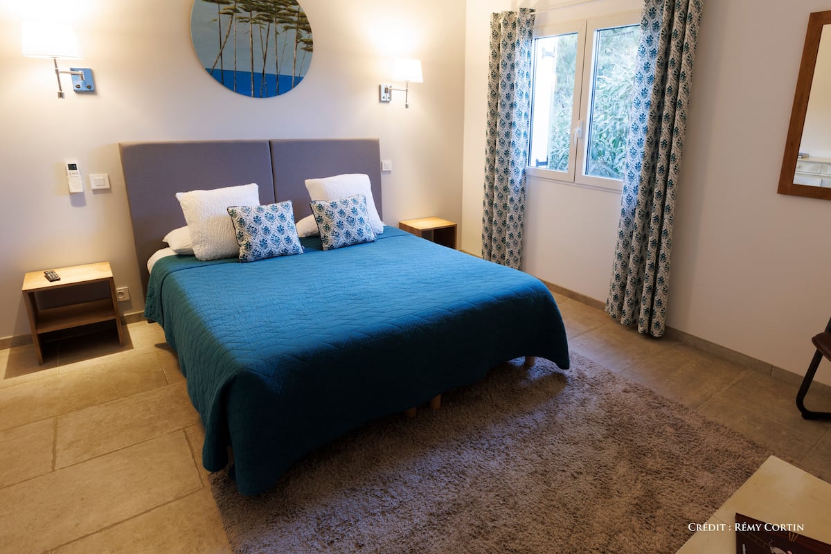 Un dormitorio con una cama grande, dos mesitas de noche, una lámpara de pared, un ventanal con cortinas estampadas y una obra de arte en la pared.